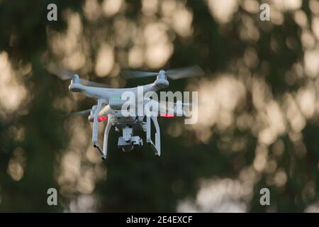 21 - Sonnenuntergang Bokeh Hintergrund hinter weißen Quadcopter Kamera Drohne. Golden Hour Sonnenlicht schafft verträumte glatte Bokeh Hintergrund scheint durch Bäume Stockfoto
