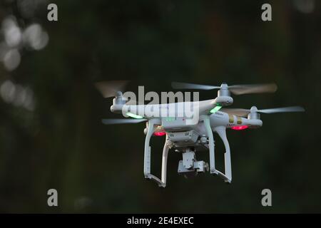 23 - Grüne und rote Orientierungslichter auf weißem Quadcopter Kamera Drohne, wie es in Richtung Dunkelheit fliegt unscharf Bäume Stockfoto