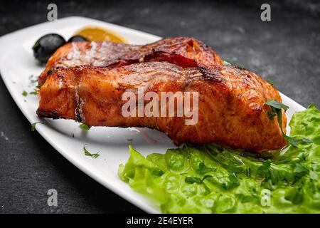 Zwei gebackene gegrillte Lachsfische auf einem weißen Teller mit Salat, Oliven und Zitrone auf dunklem Grund Stockfoto