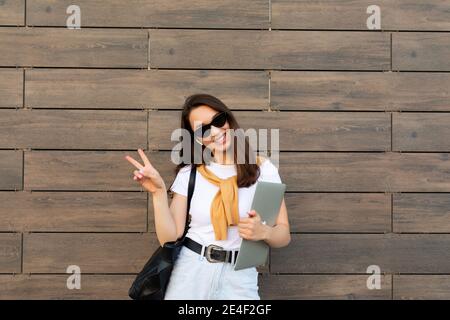 Faszinierend schön lächelnd charmant junge Brunet Mädchen Blick auf Kamera Laptop und Sonnenbrille halten, die eine Friedensgeste in Weiß zeigen t Stockfoto