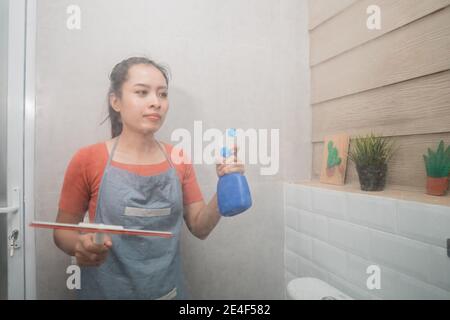 Asiatische Frau hält Flasche Spray und Scheibenwischer während der Reinigung Das Toilettenglas im Badezimmer Stockfoto