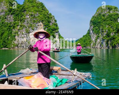 Lokale Führer in typischen violetten Uniformen und konischen Hüten nehmen Touristen mit auf eine Bootsfahrt durch die majestätischen Kalksteinberge von Halong Bay, Vietnam Stockfoto