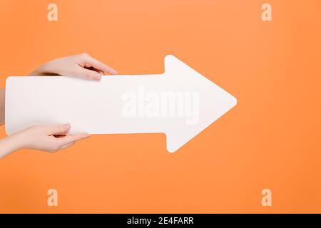 Weibliche Hände halten großen weißen Papier Pfeil, isoliert auf pastellorange Studio-Hintergrund mit Kopieplatz für Werbung. Werbebereich, Mockup Stockfoto