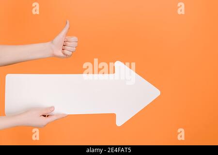 Nahaufnahme der weiblichen Hand hält großen Papierpfeil und macht Geste Daumen nach oben Geste, zeigt Zustimmung oder Zustimmung, isoliert auf pastellorange Stockfoto