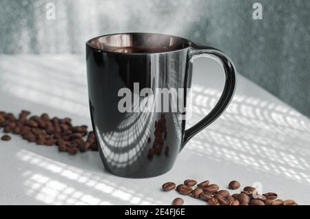 Heißer Kaffee in einer Tasse, vor dem Hintergrund von durchbrochenen Schatten aus dem Fenster und verstreuten Kaffeebohnen. Stockfoto