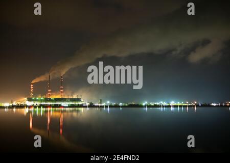 Hell erleuchtetes Kohlekraftwerk hohe Rohre mit schwarzem Rauch, der nach oben die verschmutzende Atmosphäre in der Nacht mit den Reflexen von ihm im Seewasser bewegt. Stockfoto