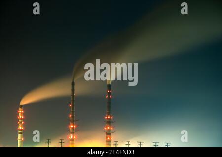 Hell erleuchtetes Kohlekraftwerk hohe Rohre mit schwarzem Rauch, der nach oben die verschmutzende Atmosphäre in der Nacht mit den Reflexen von ihm im Seewasser bewegt. Stockfoto