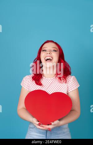 Lachende, fröhliche, überfröhliche junge Frau mit rotem Haar hält ein rotes Papierherz in zwei Händen, isoliert auf blauem Hintergrund Stockfoto