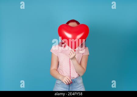 Porträt einer Frau mit roten Haaren, die sich hinter einem roten fliegenden Ballon in Form eines Herzens versteckt, isoliert auf blauem Hintergrund Stockfoto