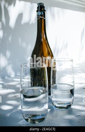 Eine Flasche und zwei Gläser Wasser stehen auf einem Weißer Rand gegen eine helle weiße Wand mit Schatten Blätter an einem sonnigen Sommertag Stockfoto