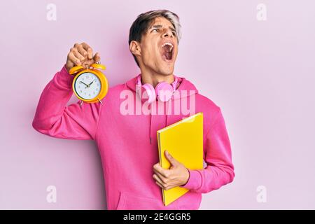 Junger hispanischer Mann hält Buch und Wecker wütend und wütend schreien frustriert und wütend, schreien mit Wut nach oben. Stockfoto