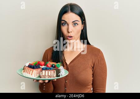 Junge Brünette Frau hält Teller mit Kuchen Scheiben erschrocken und erstaunt mit offenem Mund für Überraschung, Unglauben Gesicht Stockfoto