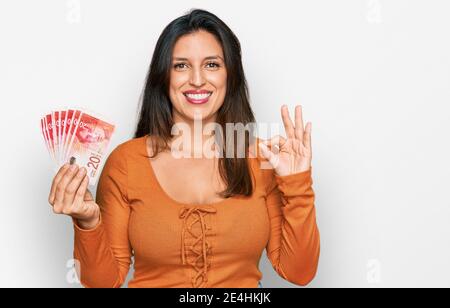 Schöne hispanische Frau hält 20 israel Schekel Banknoten tun ok Zeichen mit Fingern, lächelnd freundlich gestikulieren ausgezeichnetes Symbol Stockfoto