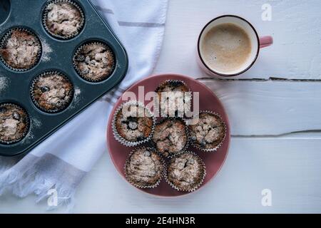 Gesunde Haferflocken-Muffins auf rosa Teller und Cappuccino-Tasse auf weißem Holztisch, Draufsicht, flach liegend. Stockfoto