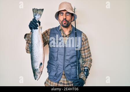 Gutaussehender hispanischer Mann mit Bart, der Angelrute hält und rohen Lachs, der Fisch ins Gesicht mit Mund macht und Augen schielt, verrückt und komisch. Stockfoto