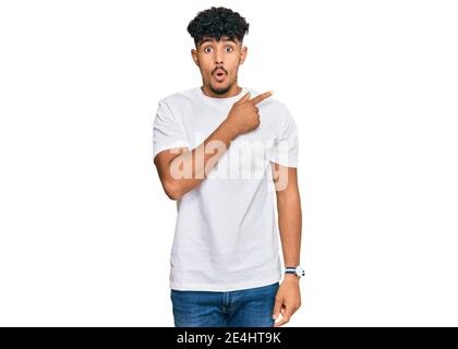 Junger arabischer Mann trägt casual weißes T-Shirt überrascht zeigt mit Finger zur Seite, offenen Mund erstaunt Ausdruck. Stockfoto