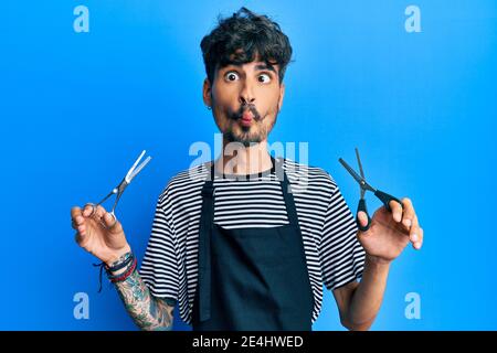 Junger hispanischer Mann trägt Friseurschürze hält Schere machen Fisch Gesicht mit Mund und schielende Augen, verrückt und komisch. Stockfoto