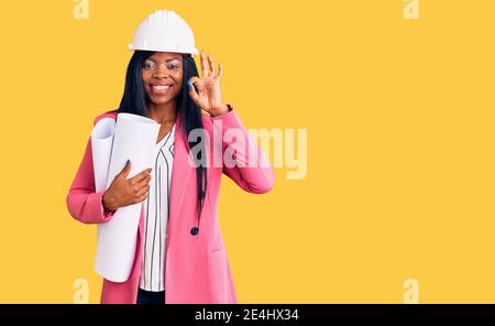 Junge afroamerikanische Frau trägt Architekt Hardhat hält Blaupausen tun ok Zeichen mit Fingern, lächelnd freundlich gestikulieren ausgezeichnete Symbol Stockfoto
