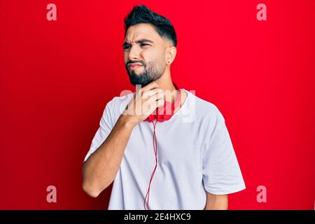 Junger Mann mit Bart, der Musik hört, mit Kopfhörern, die schmerzhaften Hals, Halsschmerzen wegen Grippe, Blutgerinnsel und Infektion berühren Stockfoto