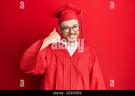 Junger Rotschopf Mann trägt rote Graduierung Kappe und Zeremonie Robe lächelnd tun Telefon Geste mit Hand und Fingern wie am Telefon zu sprechen. Commun Stockfoto