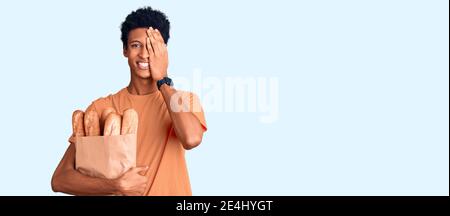 Junger afroamerikanischer Mann hält Papiertüte mit Brot, das ein Auge mit der Hand bedeckt, selbstbewusstes Lächeln im Gesicht und Überraschungsemotion. Stockfoto