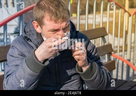 Ein kranker und schlecht aussehender Mann in einem medizinischen Anti-Virus Mask versucht, eine Zigarette an einem Bahnhof anzuzünden Plattformbank Stockfoto