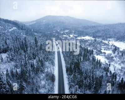 Verschneite Straße durch eine bewaldete Berglandschaft im Winter. Weihnachtsbaum Wald und Berggipfel. Stockfoto