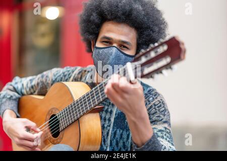 Junger Mann trägt Gesichtsmaske Gitarre spielen, während im Freien sitzen Stockfoto