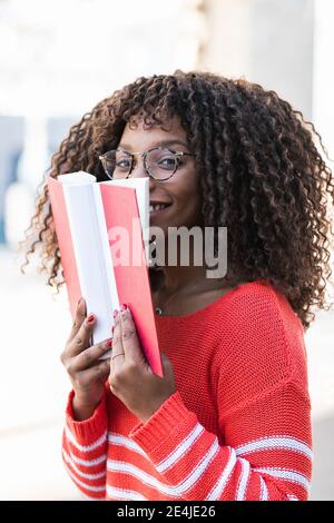 Glückliche junge Frau in warmer Kleidung, die Gesicht mit Buch bedeckt Stockfoto