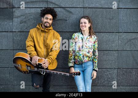 Lächelnder Mann hält .guitar, während er bei einer Frau gegen Beton steht Wand Stockfoto