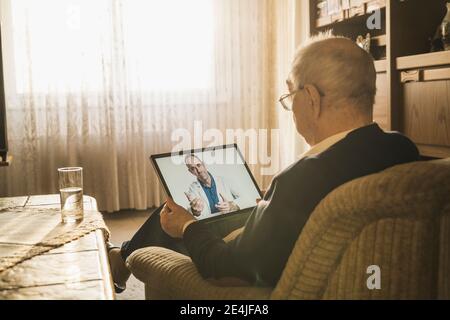 Älterer Mann, der Rat von einem männlichen Arzt bei einem Videoanruf einnahm Während Sie zu Hause auf dem Sofa sitzen Stockfoto