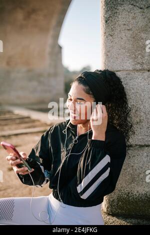 Glückliche Sportlerin, die in-Ear-Kopfhörer während der Verwendung von Mobiltelefonen justiert An sonnigen Tag Stockfoto