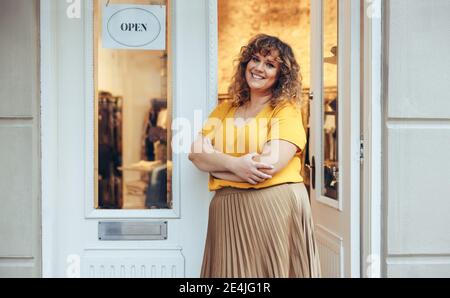 Porträt der reifen weiblichen Ladenbesitzerin lächelnd, während die Arme an der Tür ihres Modehauses gekreuzt stehen. Frau, die an der Eingangstür von ihr steht Stockfoto