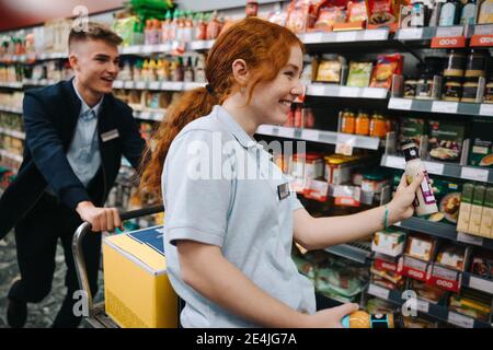 Supermarktmitarbeiter haben Spaß bei der Arbeit. Mann schiebt Frau auf Wagen, während Frau Wiederbefüllung der Regale im Lebensmittelgeschäft. Stockfoto