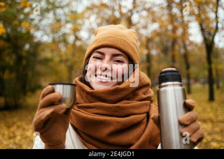 Lächelnde junge Frau mit isoliertem Getränkebehälter im Herbstpark Stockfoto