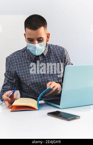 Junger Geschäftsmann mit einem Laptop, der Tagebuch auf dem Schreibtisch gegen Weiß überprüft Wand während COVID-19 Stockfoto