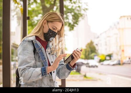 Frau mit Handy an der Bushaltestelle in der Stadt während COVID-19 Stockfoto