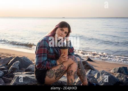 Hipster Frau mit Hand im Haar hält wiederverwendbare Kaffeetasse Beim Sitzen auf Felsen am Strand Stockfoto