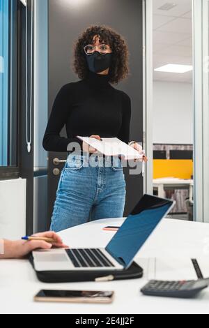 Geschäftsfrau mit Tagebuch schaut weg, während Kollege mit Laptop an Schreibtisch im Büro Stockfoto