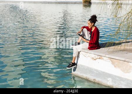Junge Frau mit digitalen Tablet, während sitzen am Rande der Promenade über den Fluss an sonnigen Tag Stockfoto