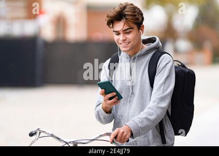 Lächelnder, modischer junger Mann, der Smartphone benutzt, während er Fahrrad rollt In der Stadt Stockfoto