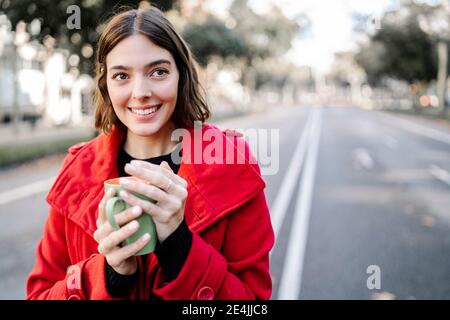 Lächelnde Frau in Winterjacke Tag träumen, während Kaffee halten becher auf der Straße Stockfoto