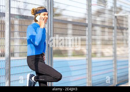 Lächelnder männlicher Athlet auf Telefonanruf gegen Zaun auf sonnig Tag Stockfoto