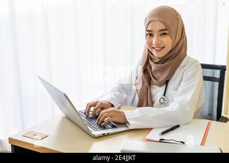 Selbstbewusste muslimische Frau Arzt am Schreibtisch sitzen, arbeiten mit Laptop-Computer in Klinik Krankenhaus Büro tippen; Blick auf die Kamera Stockfoto