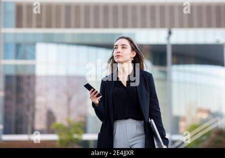 Junge Geschäftsfrau mit Laptop mit Mobiltelefon, während Sie im Freien gehen Stockfoto
