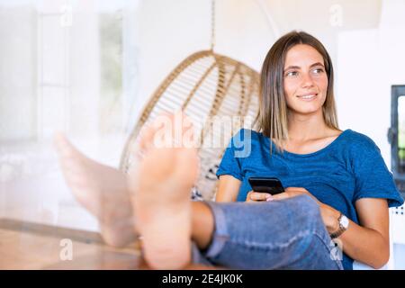 Lächelnd entspannt weibliche professionelle wegschauen, während mit smart sitzen Telefon und Füße am Tisch im Heimbüro Stockfoto