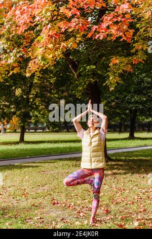 Reife Frau balanciert auf einem Bein und praktiziert Yoga im Herbstpark an sonnigen Tagen Stockfoto