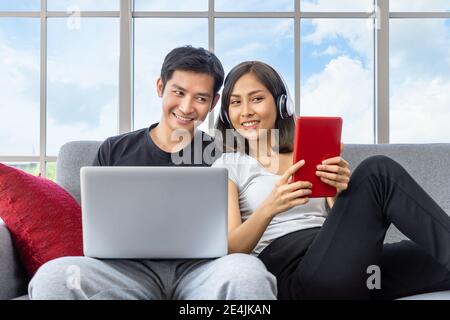 Junges asiatisches Paar entspannt sitzen auf dem Sofa mit Laptop-Computer Und ein digitales Tablet, das von zu Hause aus funktioniert Stockfoto