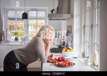 Nachdenkliche pensionierte Frau, die sich mit Erdbeeren auf der Küchentheke lehnte Wohnung Stockfoto