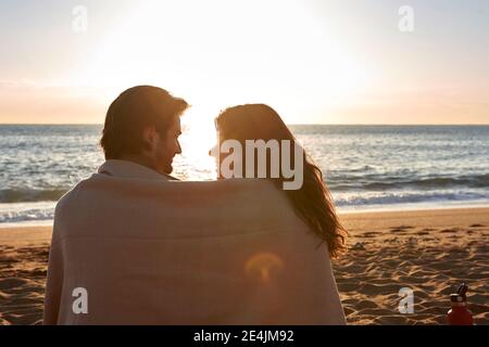 Lächelndes Paar, bedeckt mit einer Decke, während es sich ansieht Am Strand sitzen Stockfoto